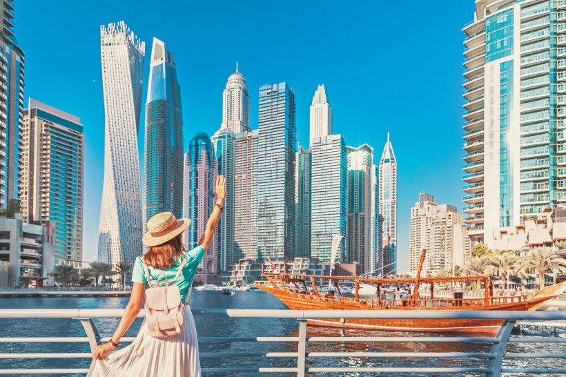 Eine Frau mit Strohhut, Rucksack und hellen Sommerkleidern steht an der Promenade der Dubai Marina. Sie blickt auf die Skyline aus modernen Hochhäusern und winkt einem traditionellen Holzboot (Dhau), das über das Wasser fährt. Der Himmel ist klar und stra