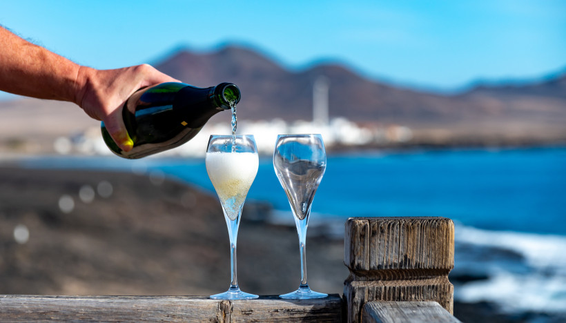 Sektgenuss mit Meerblick Sekt wird in ein Glas gegossen, daneben ein weiteres leeres Glas, im Hintergrund blaues Meer, Küste mit weißen Häusern und Vulkanbergen