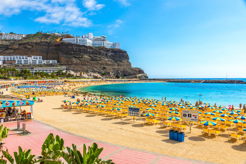 Strand von Puerto Rico auf Gran Canaria mit Liegen und blau-gelben Sonnenschirmen, Bucht und Hotelanlage am Felsen