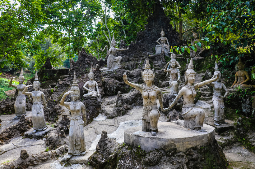 Thailand - Koh Samui Das Bild zeigt eine Sammlung kunstvoll gearbeiteter thailändischer Steinskulpturen, die in einer felsigen, bewaldeten Umgebung aufgestellt sind. Die Figuren stellen traditionelle Tänzerinnen und mythologische Wesen dar, jede in einer anderen Haltung – ste