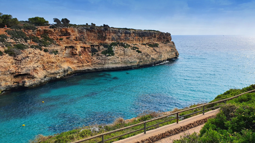 Begehbarer Küstenweg mit Geländer oberhalb der Steilküste von Calas de Mallorca mit Blick auf das offene Meer.