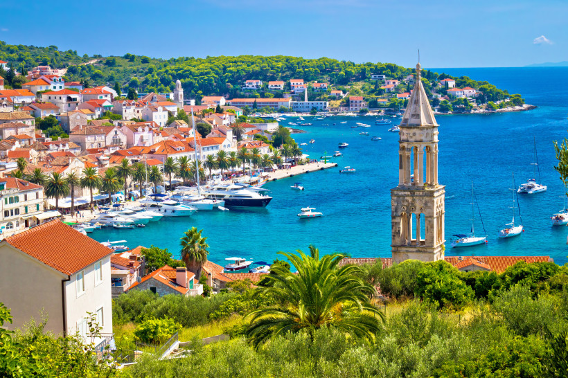 Blick auf den malerischen Hafen von Hvar-Stadt auf der kroatischen Insel Hvar. Entlang der Uferpromenade säumen Palmen die Küste, davor liegen zahlreiche Yachten und Boote im türkisblauen Wasser. Im Vordergrund ragt der historische Glockenturm einer Kirch