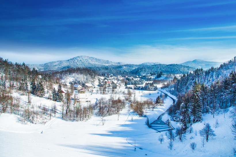 Kroatien Winterliche Landschaft mit schneebedeckten Hügeln, einem kleinen Dorf mit verstreuten Häusern und einer von Schnee gesäumten Straße, die sich durch das Tal schlängelt. Im Hintergrund erstrecken sich bewaldete Berge, deren Bäume ebenfalls mit Schnee bedeck