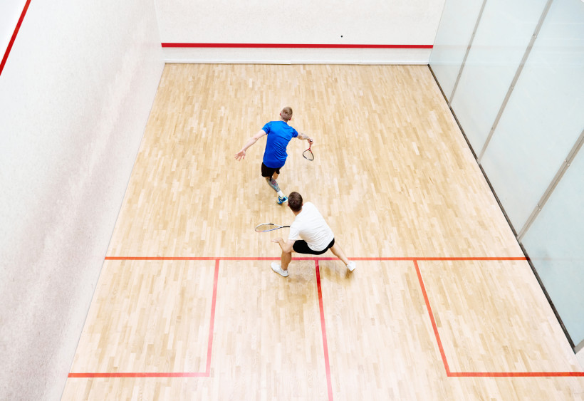 Ägypten - Marsa Alam Zwei Männer spielen Squash auf einem hellen Holzplatz. Der eine trägt ein blaues Shirt und schlägt den Ball, während der andere in einem weißen Shirt bereitsteht. Das Spielfeld ist von roten Linien begrenzt.