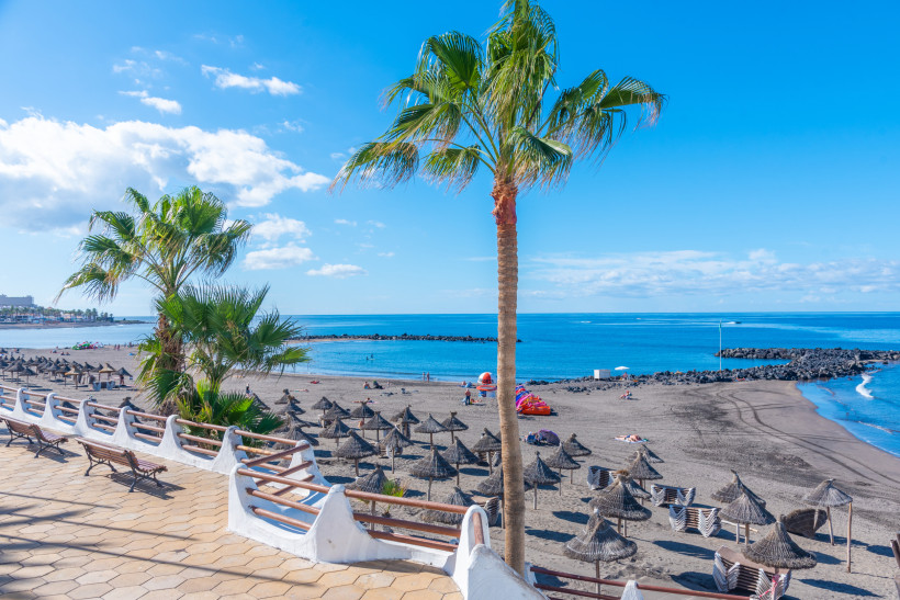 Teneriffa - Playa de Troya Playa de Troya in Costa Adeje, Teneriffa: Strand mit dunklem Vulkansand, Palmen, Sonnenschirmen und ruhigem Wasser durch Wellenbrecher an der Promenade.