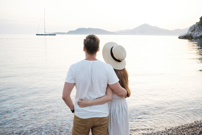 Mallorca Ein verliebtes Paar steht Arm in Arm am Strand und blickt auf das Meer bei Sonnenuntergang. Im Hintergrund ist ein Segelboot zu sehen