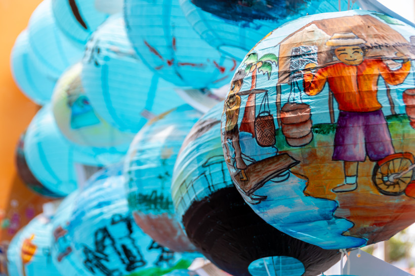 Singapur Mehrere hellblaue Papierlaternen mit bunten handgemalten Motiven, darunter eine Zeichnung eines lächelnden Mannes mit Strohhut vor einer Hütte. Laternen hängen eng beieinander und vermitteln eine festliche Stimmung.