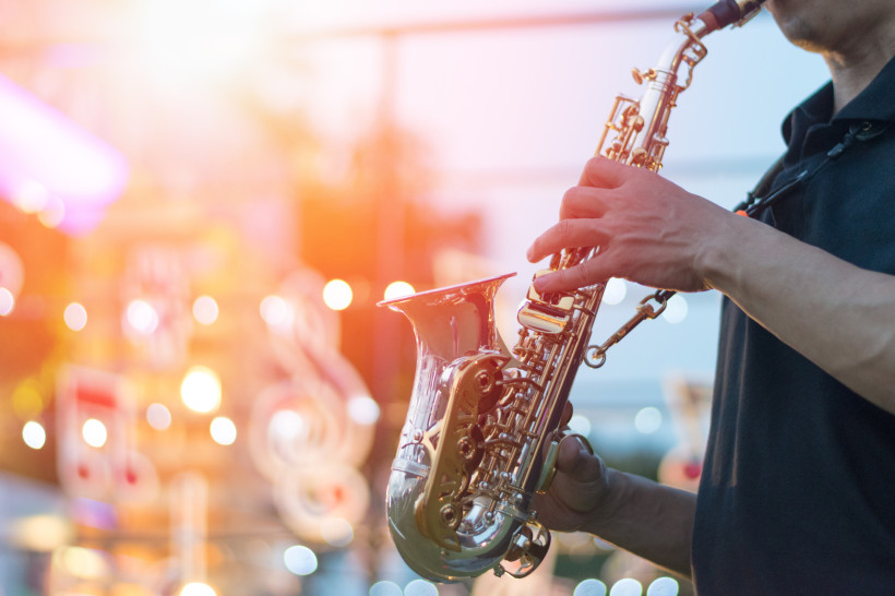 Das Bild zeigt einen Musiker, der ein glänzendes Saxofon spielt. Die Szene ist stimmungsvoll beleuchtet, mit warmem Licht und verschwommenen Musiknoten im Hintergrund – es wirkt wie ein Konzert oder ein Musikfestival bei Sonnenuntergang.