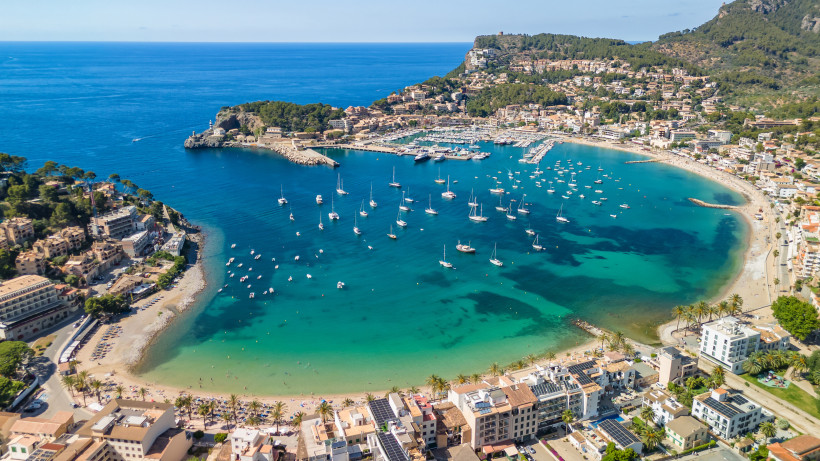 Mallorca - Port de Sóller Eine lebhafte Küstenstadt mit vielen weißen Segelbooten im türkisfarbenen Wasser. Die Stadt liegt in einer malerischen Bucht, umgeben von grünen Hügeln und Bergen. Im Hintergrund erstreckt sich eine beeindruckende Gebirgskette.