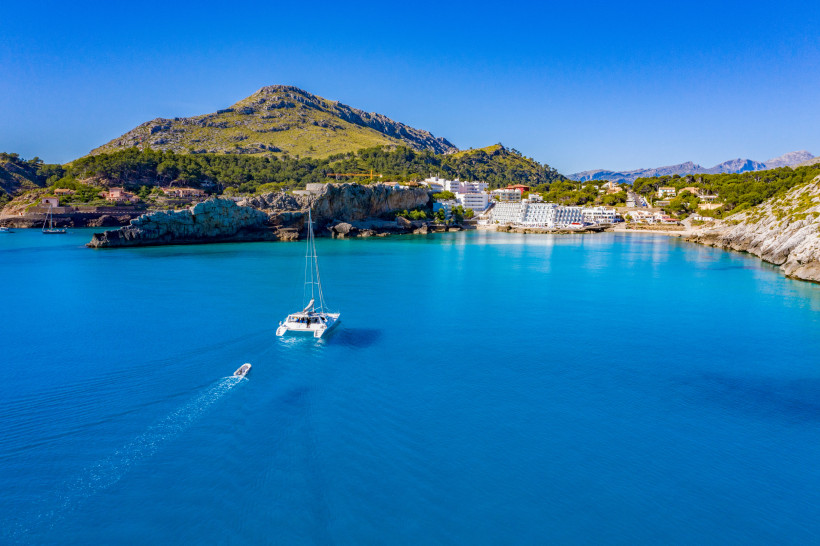 Bucht von Cala San Vicente mit türkisfarbenem Wasser, Segelboot vor der Küste und Felslandschaft im Hintergrund.