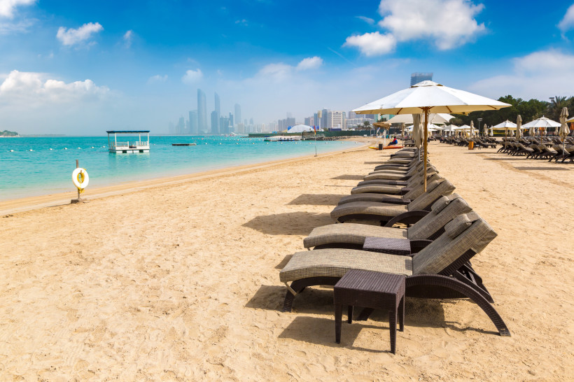 Luxuriöser Sandstrand in Abu Dhabi mit Liegestühlen und Sonnenschirmen, mit Blick auf die Skyline der Stadt im Hintergrund