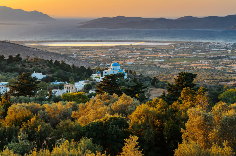Kos Malerischer Sonnenuntergang über griechischem Dorf mit weißer Kirche und blauer Kuppel, umgeben von grüner Hügellandschaft, Olivenhainen und dem Meer in der Ferne.