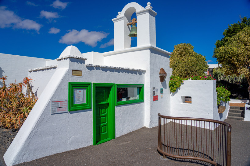 Eingang der Fundación César Manrique – Traditionelle Architektur Lanzarotes Weißes Gebäude mit grünem Eingang und Glockenturm am Eingang der Fundación César Manrique in Tahíche auf Lanzarote, umgeben von Pflanzen und blauem Himmel.