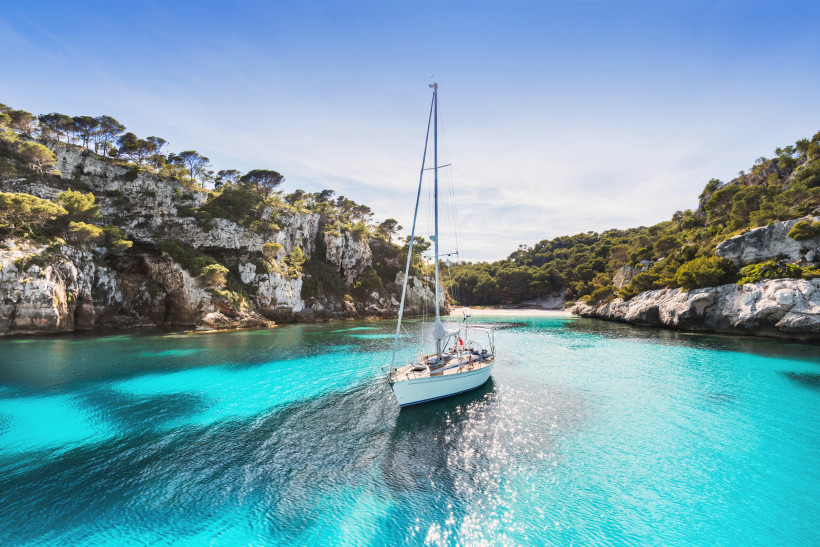 Ibiza Ein einsames Segelboot gleitet durch kristallklares, türkisfarbenes Wasser in einer malerischen Bucht. Umgeben ist die Szene von felsigen Klippen und üppigem Pinienwald. Die Sonne spiegelt sich glitzernd auf dem Wasser, der Himmel ist strahlend blau – ein