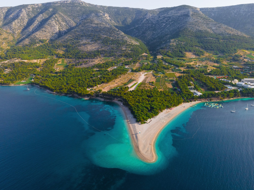 Luftaufnahme des bekannten Zlatni Rat Strandes auf der kroatischen Insel Brač. Die landzungeförmige Kiesbank ragt sichelförmig ins türkisfarbene Meer. Umgeben ist der Strand von dichten Pinienwäldern und einer bergigen Küstenlandschaft im Hintergrund. Ein