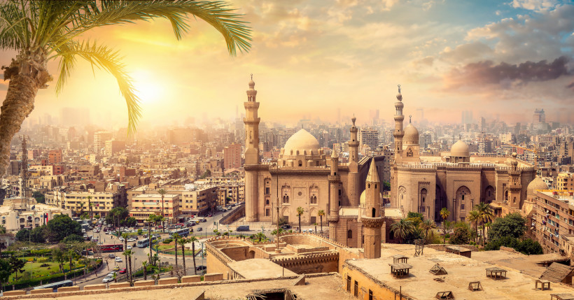 Das Bild zeigt ein weitläufiges Panorama der ägyptischen Hauptstadt Kairo bei Sonnenuntergang. Im Vordergrund steht die eindrucksvolle Sultan-Hassan-Moschee mit ihrer großen Kuppel und mehreren Minaretten, umgeben von Palmen und historischen Mauern. Die m