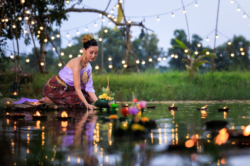 Thailand - Koh Samui Das Bild zeigt eine Frau in prachtvoller traditioneller thailändischer Kleidung, die in ruhiger Haltung ein mit Blumen und Kerze geschmücktes Krathong auf einem Teich platziert. Es ist Dämmerung, im Hintergrund leuchten warme Lichterketten über der Wiese