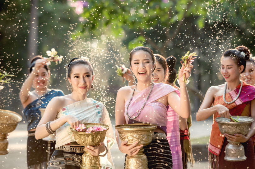 Thailand - Koh Samui Das Bild zeigt eine fröhliche Gruppe junger Frauen in traditioneller thailändischer Kleidung während einer Feier im Freien. Sie tragen elegante Seidengewänder in kräftigen Farben und halten vergoldete Schalen, gefüllt mit Blumen und Wasser. In der Luft sp