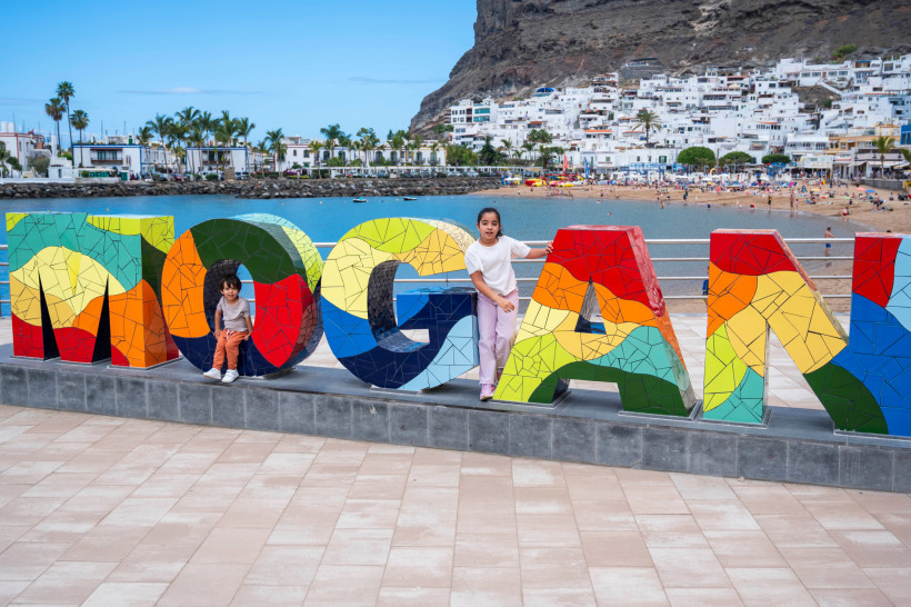 Puerto de Mogán: Promenade mit „MOGAN“-Skulptur, Strand und Häusern am Hang Bunte Buchstaben-Skulptur „MOGAN“ an der Promenade von Puerto de Mogán, im Hintergrund Strand und weiße Häuser am Hang