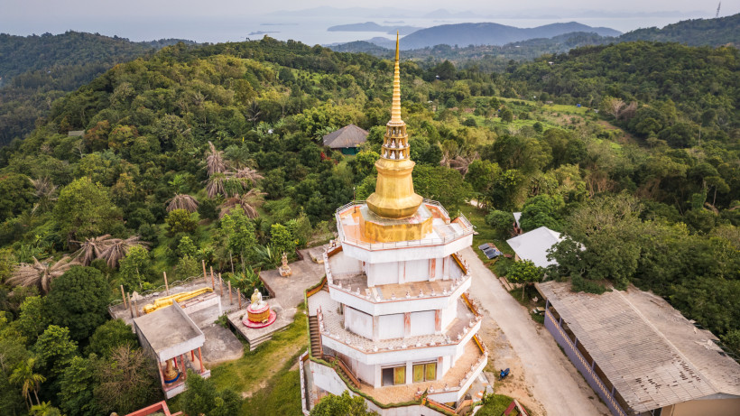Thailand - Koh Samui Das Bild zeigt eine beeindruckende Luftaufnahme eines buddhistischen Tempels mit einer goldenen, mehrstufigen Turmspitze. Der Tempel ist achteckig gebaut, weiß mit goldenen und roten Verzierungen, und thront auf einem bewaldeten Hügel. Rundherum erstreckt