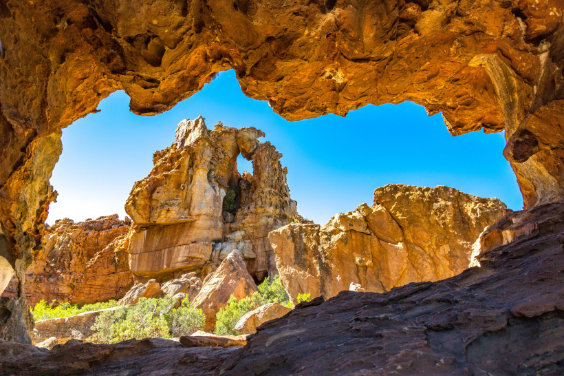 Südafrika Blick aus einer natürlichen Felsöffnung auf bizarre, rot-orange Felsformationen unter strahlend blauem Himmel. Die Szene zeigt eine beeindruckende Landschaft mit zerklüfteten Gesteinsformationen und grünem Buschwerk im Vordergrund, typisch für die Cederbe