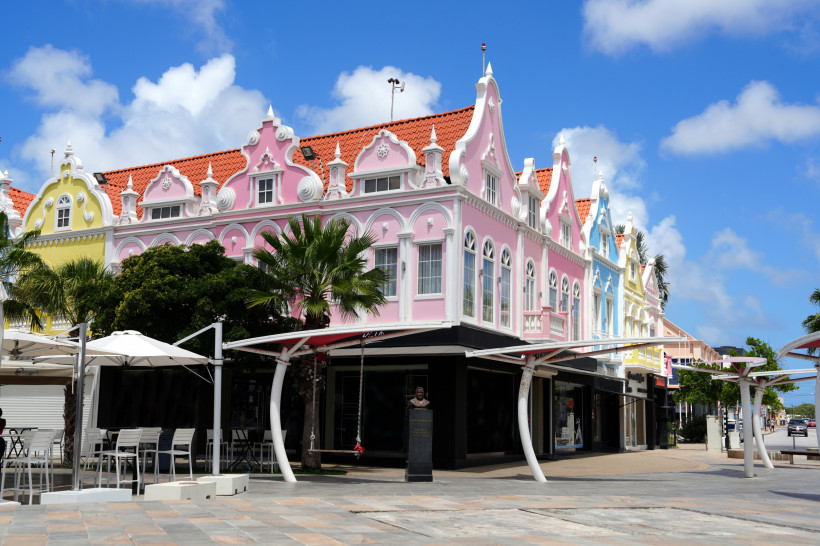 Reihe farbenfroher, kolonial inspirierter Gebäude in Oranjestad, Aruba. Die Fassaden sind in Rosa, Gelb und Blau gestrichen, mit weißen Ornamenten und roten Ziegeldächern. Palmen und weiße Sonnenschirme ergänzen das Straßenbild.