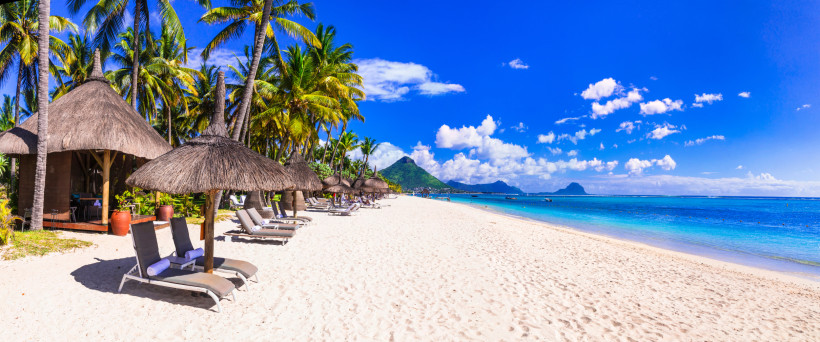 Mauritius traumhafte Urlaubskulisse an einem tropischen Sandstrand. Im Vordergrund stehen luxuriöse Sonnenliegen unter Strohdach-Sonnenschirmen, die Schatten spenden und für eine entspannte Atmosphäre sorgen. Umgeben von üppigen, hohen Palmen vermittelt der Strand