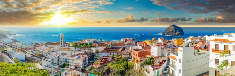 Panoramablick auf Garachico und den Atlantik bei Sonnenuntergang Panorama von Garachico auf Teneriffa mit hellen Häusern und roten Dächern, der markanten Kirche und dem Atlantik im Hintergrund; links der Küstenverlauf, rechts der kleine Felseninsel „Roque de Garachico“, beleuchtet von warmem Sonnenuntergangslicht.
