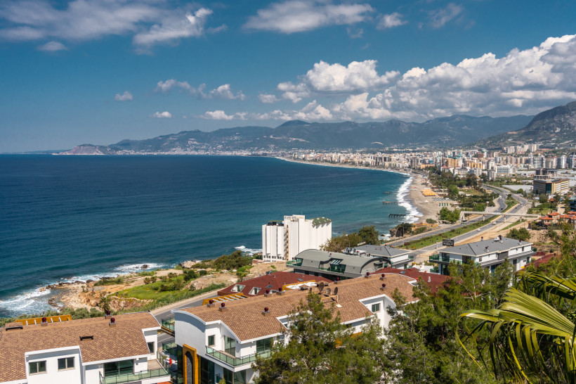 Das Bild zeigt einen weiten Blick auf die Ostküste von Alanya. Im Vordergrund befinden sich moderne Wohnanlagen mit Flachdächern. Entlang der Küste verläuft eine breite Straße und zahlreiche Hotel- und Wohngebäude. Im Hintergrund sind das Mittelmeer, der 