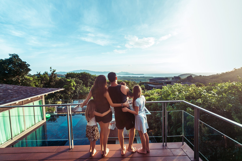 Thailand - Phuket Eine Familie mit zwei Erwachsenen und drei Kindern steht barfuß auf einer modernen Terrasse mit Glasgeländer. Sie schauen gemeinsam in Richtung einer üppig grünen Landschaft mit Bäumen, vereinzelten Gebäuden und einem Blick bis zum Meer oder einem See in