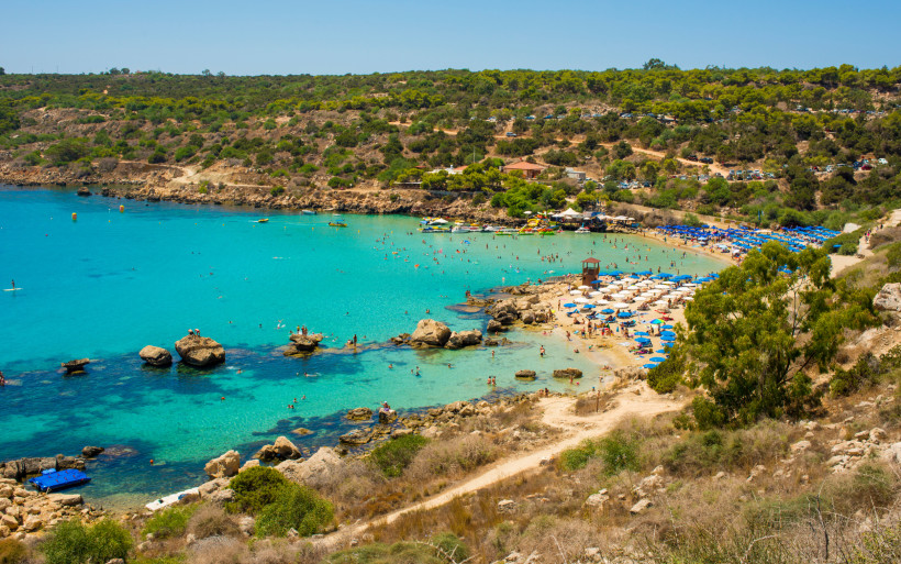 Konnos Bay bei Agia Napa/Protaras: geschützte Sandbucht mit türkisfarbenem Wasser, Felsen und Reihen blauer Sonnenschirme.