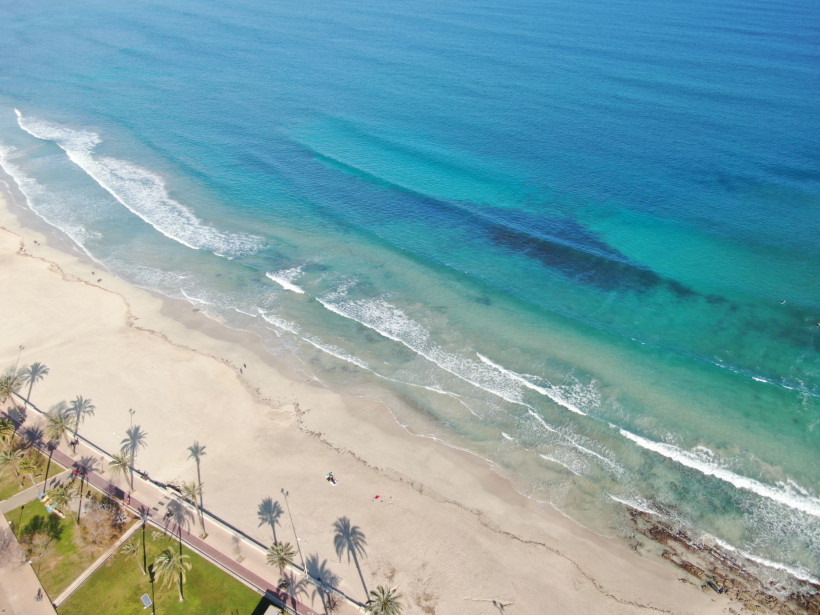 Luftaufnahme des gepflasterten Küstenwegs direkt am Strand zwischen Cala Bona und Cala Millor auf Mallorca, Palmenpromenade mit Blick auf das türkisfarbene Meer und den langen Sandstrand.