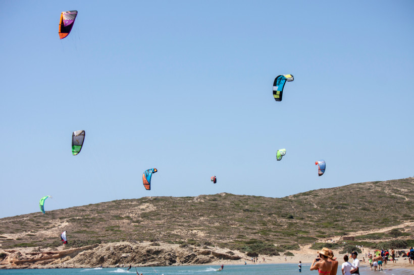 Mehrere bunte Kiteschirme beim Kitesurfen über einer türkisfarbenen Bucht, felsige Küste und hügelige Landschaft im Hintergrund