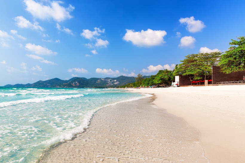 Weißer Sandstrand mit sanften Wellen und türkisblauem Wasser am Chaweng Beach auf Koh Samui, Thailand, mit grünen Bergen im Hintergrund