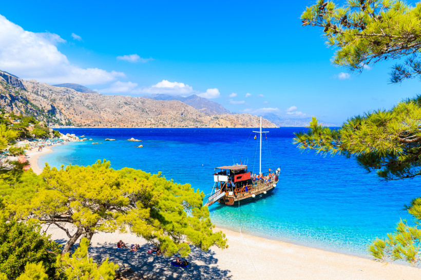 Kos Traumhafte Mittelmeerbucht mit türkisblauem Wasser, Pinienbäumen und einem Holzsegelschiff am weißen Kiesstrand vor einer felsigen Berglandschaft.
