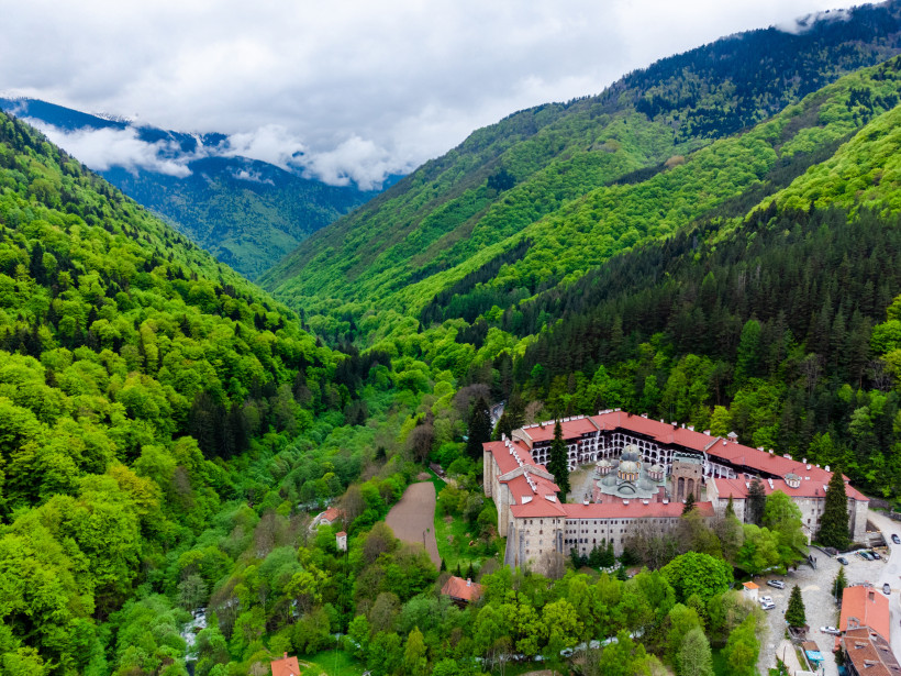 Luftaufnahme des Rila-Klosters inmitten dichter grüner Bergwälder. Die Klosteranlage mit roten Dächern und einer zentralen Kirche liegt eingebettet in einem bewaldeten Tal mit umgebenden Hügeln und Wolken am Horizont.