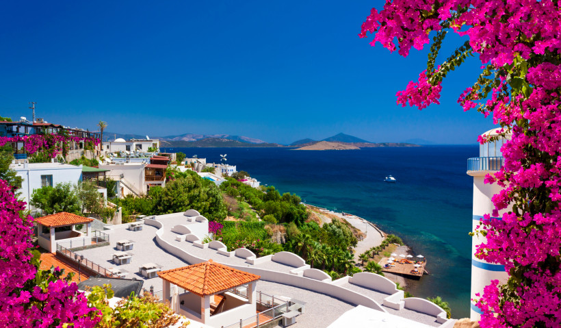 Mediterraner Küstenort mit weißen Häusern, pinken Bougainvillea und Blick auf das tiefblaue Meer