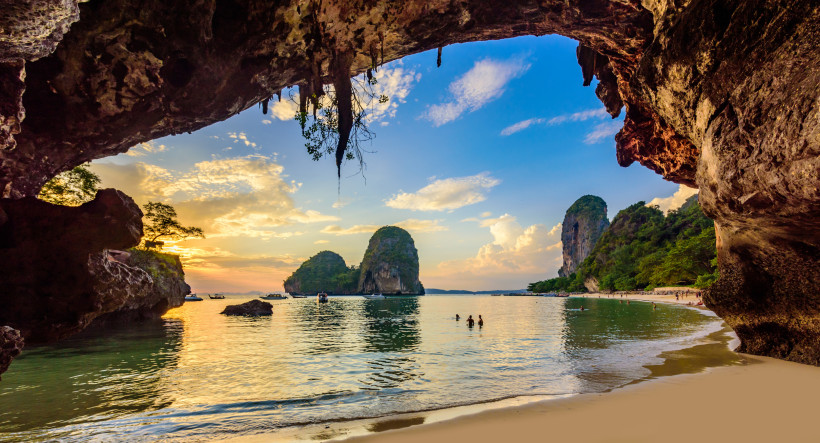 Sonnenuntergang am Phra Nang Cave Beach in Krabi: Blick aus der Höhle auf Meer, Karstfelsen und badende Urlauber.