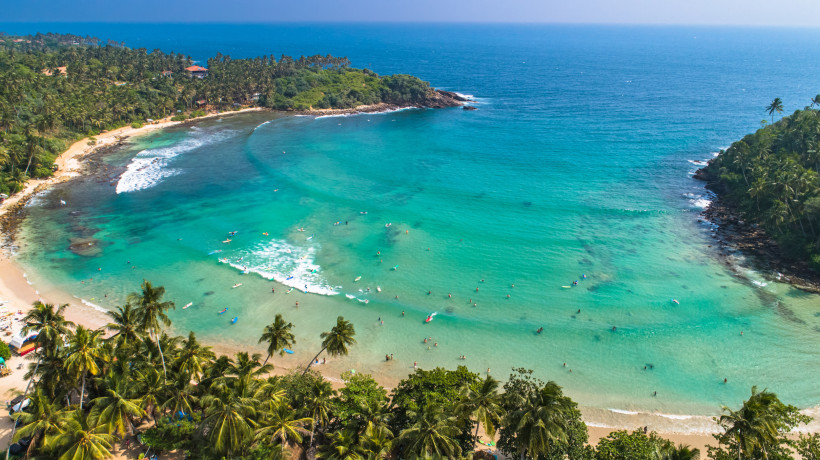 Luftaufnahme einer tropischen Bucht mit türkisblauem Wasser, feinem Sandstrand und vielen Palmen. Zahlreiche Menschen baden oder paddeln auf Surfboards im flachen Wasser. Die Bucht ist von dichter, grüner Vegetation und Felsen umgeben. Im Hintergrund erst