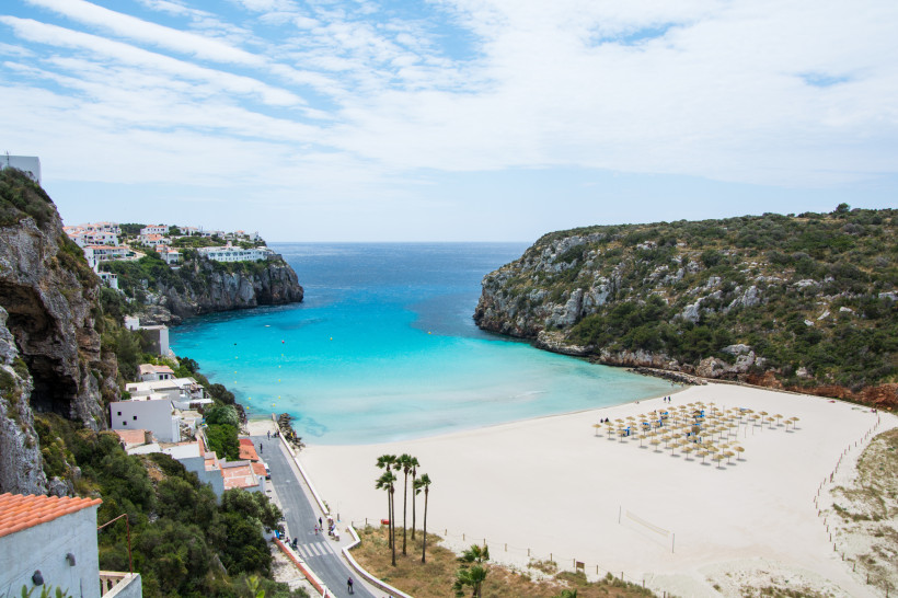 Breiter weißer Sandstrand in einer geschützten Bucht auf Menorca. Auf dem Strand stehen geordnet zahlreiche Strohmsonnenschirme. Links und rechts ragen grüne Felsklippen ins türkisfarbene Meer. Auf dem linken Hang sind weiße Häuser mit Blick aufs Meer geb