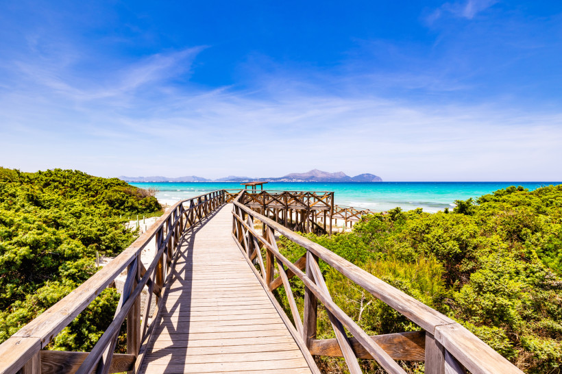 Holzsteg durch Dünen zur Bucht Es Comú mit türkisfarbenem Meer und Bergen am Horizont