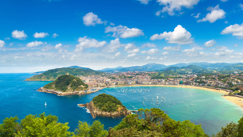 Panoramablick auf San Sebastián mit der muschelförmigen Bucht La Concha, türkisfarbenem Meer, vielen Booten und grünen Hügeln im Hintergrund.