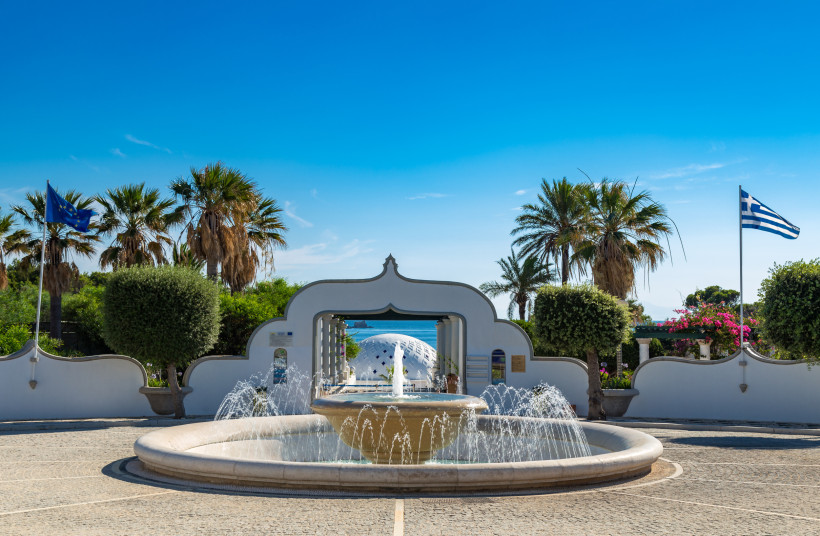 Brunnen vor weißem Torbogen mit Palmen, griechischer Flagge und Blick auf das Meer und ein kuppelförmiges Gebäude auf Rhodos