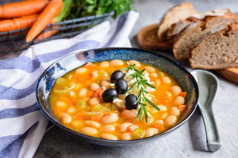 Schüssel Fasolada, griechische Bohnensuppe mit Karotten und Sellerie, garniert mit Oliven, Feta und Rosmarin; daneben Brot