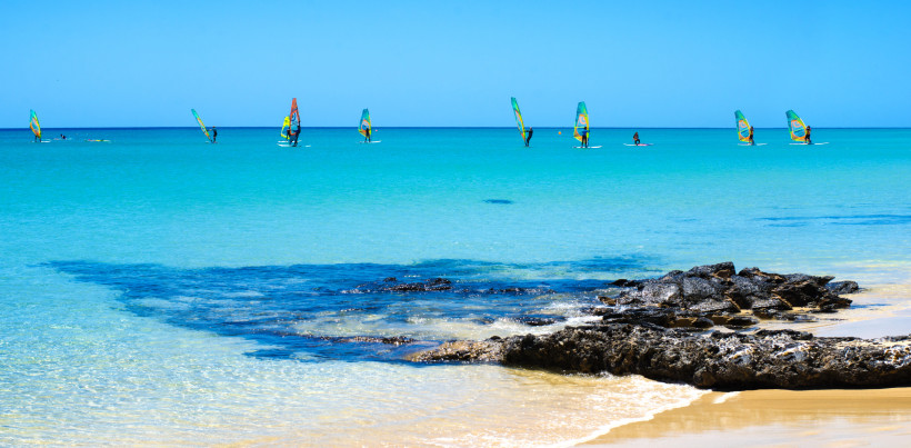 Fuerteventura Türkisfarbenes Meer mit mehreren Windsurfern, die auf dem Wasser segeln. Im Vordergrund goldener Sandstrand.