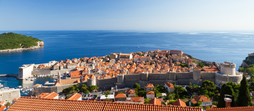 Panoramablick auf die Altstadt von Dubrovnik mit roten Ziegeldächern, die vollständig von einer Stadtmauer umgeben ist. Im Vordergrund das tiefblaue Meer und im Hintergrund eine bewaldete Insel.