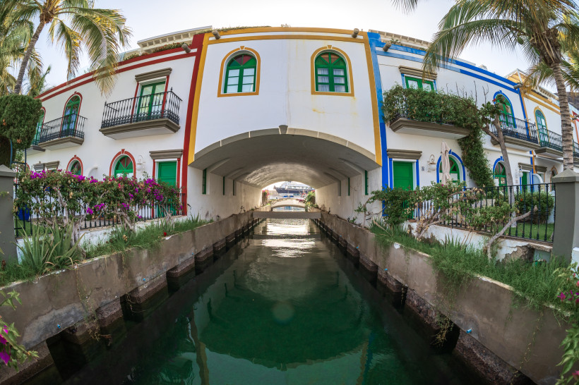 Kanäle und Brücken (Little Venice) in Puerto de Mogán Kanal in Puerto de Mogán mit Brücke unter einem weiß gestrichenen Gebäude mit bunten Fassadenstreifen, Balkonen und Palmen