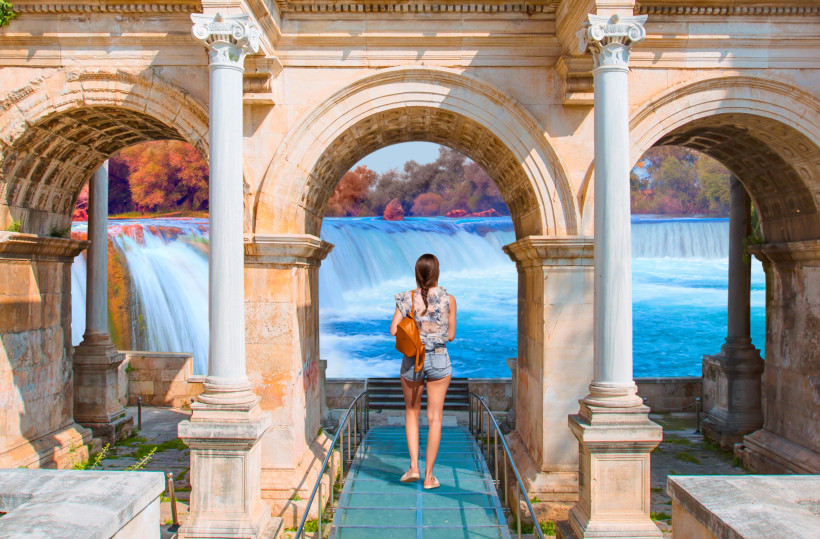 Eine Frau steht auf einem Glassteg zwischen antiken Steinbögen und blickt auf einen breiten Wasserfall mit türkisblauem Wasser. Im Hintergrund sind bunte Bäume und ein sonniger Himmel zu sehen.