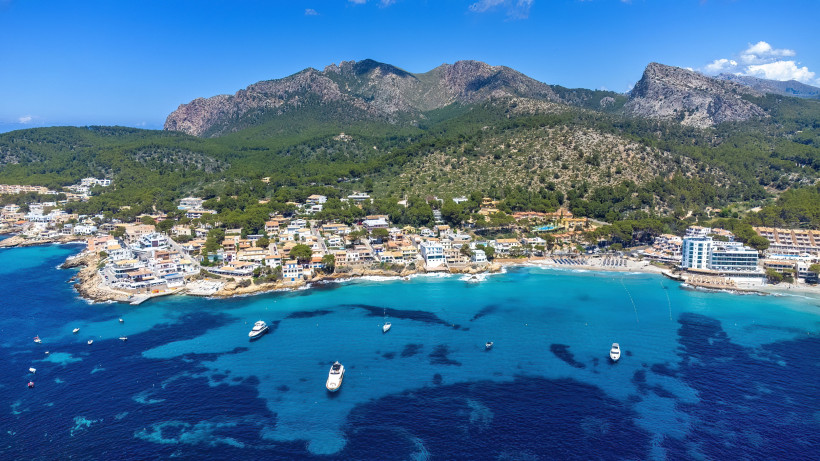 Luftaufnahme von Sant Elm mit Badebucht, Booten im Wasser und Tramuntana-Gebirge im Hintergrund