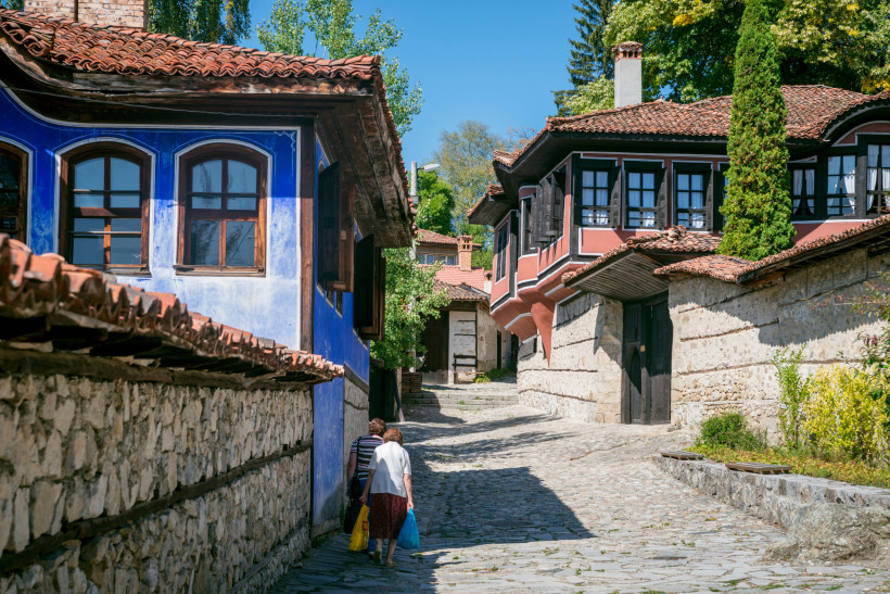 Zwei ältere Menschen gehen eine gepflasterte Gasse entlang in Koprivshtitsa. Links ein auffällig blau gestrichenes Haus mit Holzdach, rechts ein rot-schwarzes Gebäude mit traditionellen Holzfenstern. Umgeben von Steinmauern und viel Grün.